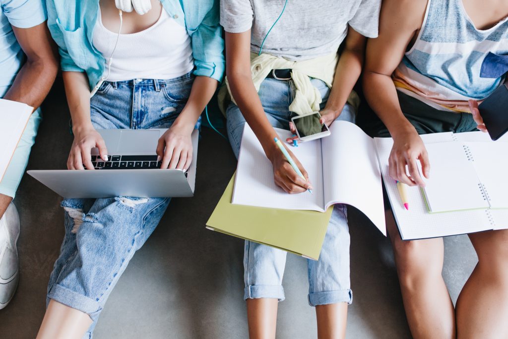 Close-up overhead portrait of girl in blue shirt and jeans holding laptop on knees while sitting beside university mates. Female student writing lecture in notebook and using phone between friends.